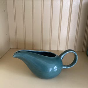 May include: A teal ceramic gravy boat with a curved spout and a rounded handle. The interior is a darker shade of teal. The gravy boat is on a light-colored surface.