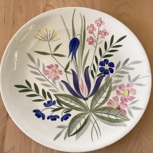 Könnte beinhalten: Ein runder, weißer Keramikteller mit floralem Design. Das Design zeigt einen Blumenstrauß in Rosa-, Blau-, Gelb- und Lilatönen mit grünen und grauen Blättern. Der Teller steht auf einer hellbraunen Holzoberfläche.