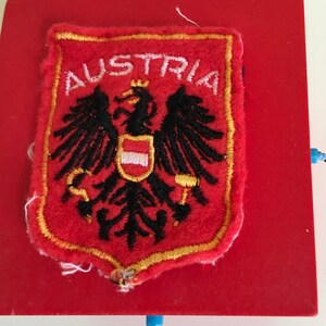 May include: A red embroidered patch featuring the Austrian coat of arms, including a black eagle with a crown and a shield. The word "AUSTRIA" is embroidered in white above the emblem. The patch has a gold border and is set on a red surface.