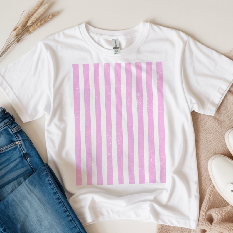 Puede incluir: Camiseta blanca con un dise&ntilde;o de rayas rosas verticales. La camiseta est&aacute; hecha de un material suave y se muestra con vaqueros azules, zapatos beige y plantas secas. La camiseta es una prenda informal.