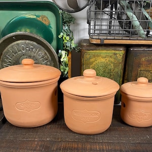 May include: Three terracotta pots with lids, labeled "Onion Pot", "Mushroom Pot", and "Garlic Pot". The pots are cylindrical with a rounded lid and a small knob on top. The pots are a light orange color.