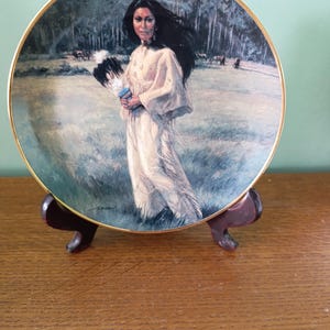 May include: Decorative plate with a gold rim, depicting a Native American woman in a white dress, holding a fan. The plate is displayed on a wooden stand. The background shows a landscape with trees and mountains.