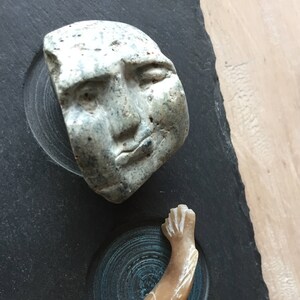 May include: A black slate board with three carved stone sculptures. The top sculpture is a face, the middle sculpture is a fish, and the bottom sculpture is a smooth, brown stone.