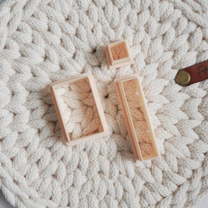 Op de afbeelding: Een crèmekleurige, ronde, geweven placemat met een gevlochten textuur. Drie lichtroze, rechthoekige koekjessnijders liggen op de placemat. Een bruin leren label met een gouden klinknagel bevindt zich aan de rechterkant.