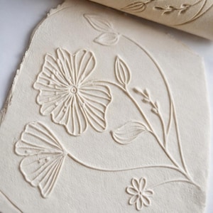 Könnte beinhalten: Eine hellbeige Tonplatte mit einem geprägten Blumendesign, das eine große Blume, kleinere Blumen, Blätter und Stiele zeigt. Ein passender Nudelholz mit dem gleichen Blumenmuster befindet sich im Hintergrund. Ideal für Töpferei und Basteln.