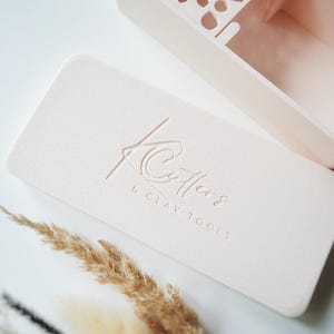 Könnte beinhalten: Eine hellrosa rechteckige Aufbewahrungsbox mit einem Deckel, der den geprägten Text K Cutters & CLAY TOOLS trägt. Der Deckel ist glatt, während die Basis auf einer Seite ein dekoratives geometrisches Ausschnittmuster zur Werkzeugaufbewahrung hat.