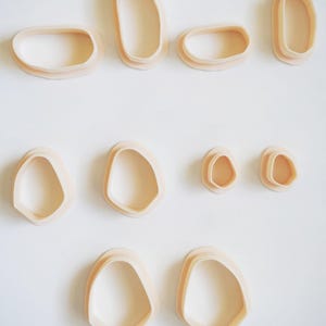 Könnte beinhalten: Zehn beige Ausstecher in organischer Form auf weißem Hintergrund. Die Ausstecher, in verschiedenen Größen, haben abgerundete Kanten und ein glattes Design. Geeignet für Back- und Bastelprojekte. Maße in cm.