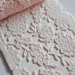 Op de afbeelding: Een lichtroze deegroller met een bloemenpatroon wordt getoond naast een bijpassend vel klei met hetzelfde reliëfontwerp. Het patroon toont gedetailleerde bloemen en bladeren, waardoor een textuuroppervlak ontstaat.
