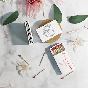 Puede incluir: Cajas de cerillas blancas con cerillas de punta roja, dispersas con cerillas sueltas y acentos florales. Una caja est&aacute; abierta, mostrando cerillas, y otra est&aacute; cerrada. Tambi&eacute;n hay una peque&ntilde;a tarjeta con una ilustraci&oacute;n en blanco y negro y la palabra "cheers!"