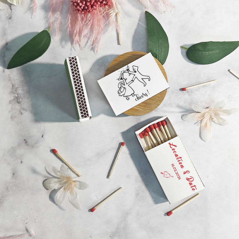 Puede incluir: Cajas de cerillas blancas con cerillas de punta roja, dispersas con cerillas sueltas y acentos florales. Una caja est&aacute; abierta, mostrando cerillas, y otra est&aacute; cerrada. Tambi&eacute;n hay una peque&ntilde;a tarjeta con una ilustraci&oacute;n en blanco y negro y la palabra "cheers!"