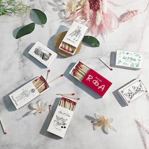 Puede incluir: Surtido de cajas de cerillas decorativas con cerillas de punta roja. Las cajas presentan varios dise&ntilde;os, incluyendo ilustraciones tem&aacute;ticas de boda, retratos de gatos y el texto "Perfect Match".