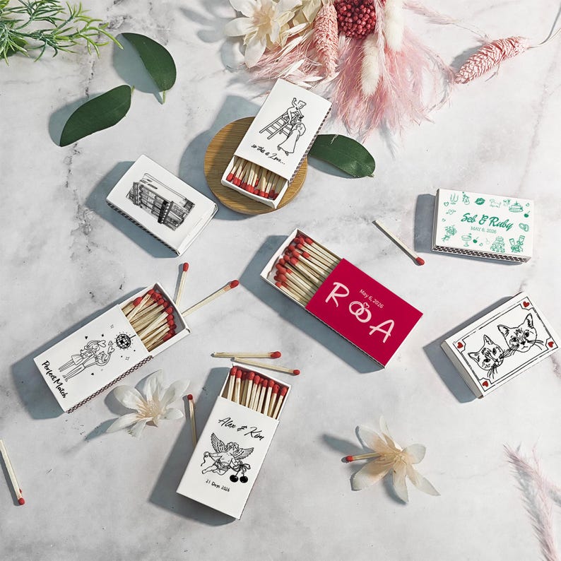 Puede incluir: Surtido de cajas de cerillas decorativas con cerillas de punta roja. Las cajas presentan varios dise&ntilde;os, incluyendo ilustraciones tem&aacute;ticas de boda, retratos de gatos y el texto "Perfect Match".