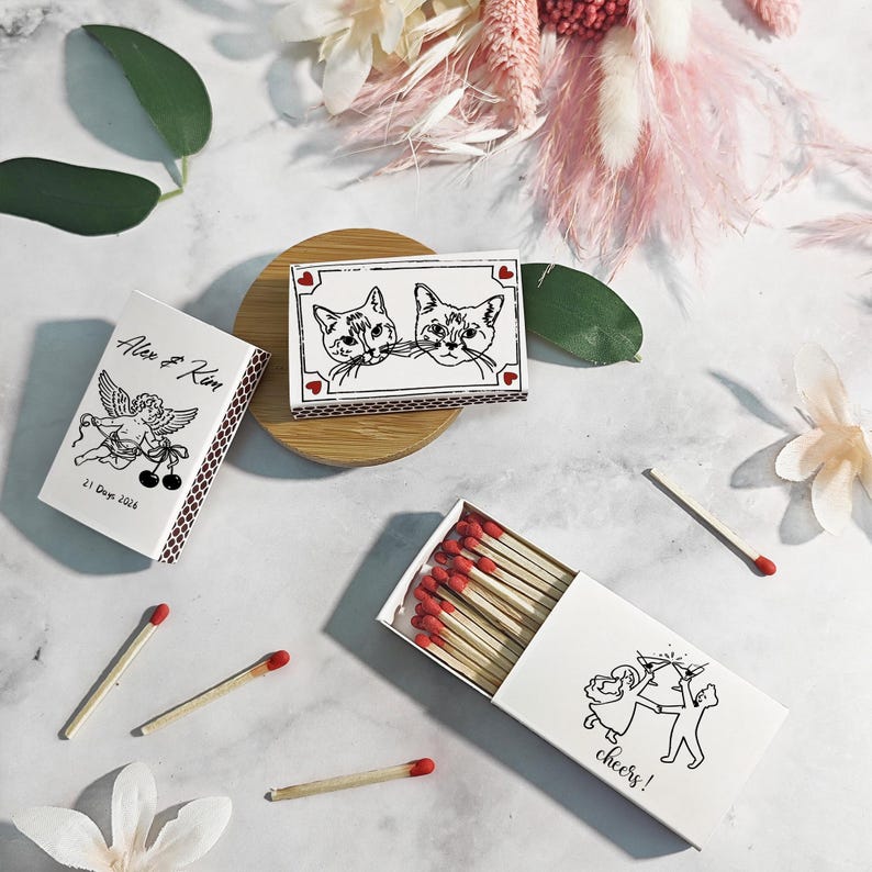 Puede incluir: Cajas de cerillas blancas con cerillas de punta roja. Una caja presenta una ilustraci&oacute;n en blanco y negro de dos gatos, otra tiene un dise&ntilde;o de querub&iacute;n con texto, y una tercera dice "cheers!" con una ilustraci&oacute;n de pareja. Las cerillas est&aacute;n dispersas.