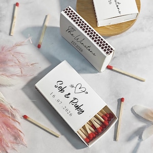 Puede incluir: Cajas de cerillas blancas con cerillas de punta roja. Una caja est&aacute; abierta, mostrando las cerillas, y otra est&aacute; parcialmente abierta. Las cajas tienen texto personalizado, incluyendo nombres y una fecha.
