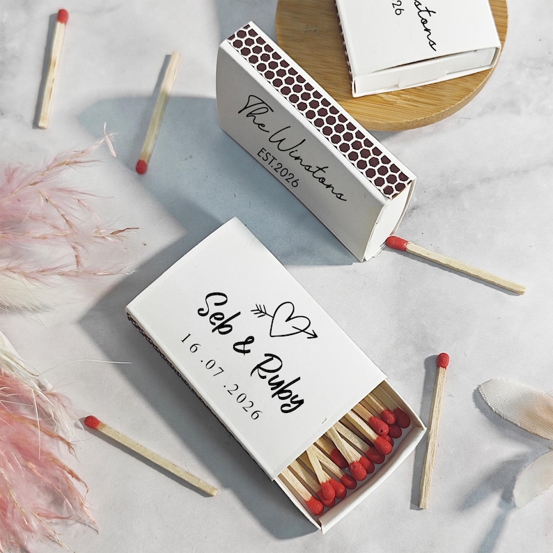 Puede incluir: Cajas de cerillas blancas con cerillas de punta roja. Una caja est&aacute; abierta, mostrando las cerillas, y otra est&aacute; parcialmente abierta. Las cajas tienen texto personalizado, incluyendo nombres y una fecha.
