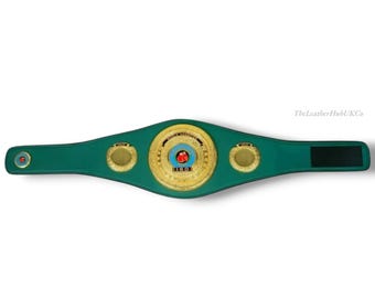 May include: A green boxing championship belt with gold accents. The belt features a large central emblem with a red jewel and the text "IBO". The belt is made of leather and has a black velcro closure.