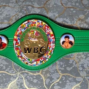 May include: A green boxing championship belt with a gold buckle and a central emblem featuring the letters "WBC". The belt has two circular portraits and a gold and multi-colored central emblem.