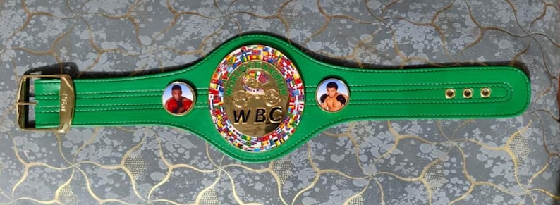 May include: A green boxing championship belt with a gold buckle and a central emblem featuring the letters "WBC". The belt has two circular portraits and a gold and multi-colored central emblem.