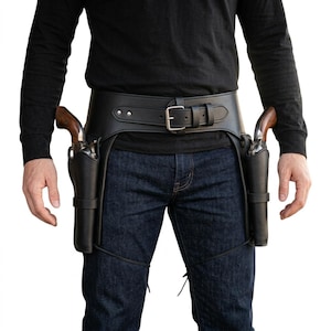 May include: A black leather gun belt featuring a wide waist strap with a silver-toned metal buckle and two attached holsters, each containing a revolver with a wooden grip. The holsters are secured to the thighs with thin black leather straps.
