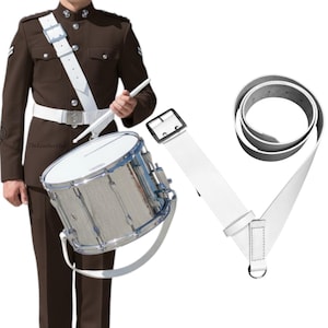 May include: A polished silver snare drum with a white strap, a white belt, and a white shoulder strap. The drum is held by a person in a brown uniform. A white belt with a black buckle is also shown.