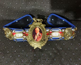 May include: A championship boxing belt with a gold central medallion featuring a portrait. The belt has blue, white, and red accents, with gold detailing and additional medallions. The belt is designed with a blue padded backing.