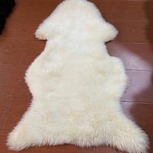 Puede incluir: Alfombra de piel de oveja color crema con una forma única. La alfombra tiene una textura suave y esponjosa y está colocada sobre un suelo de madera. La forma de la alfombra es irregular, con bordes redondeados y una forma ligeramente alargada.
