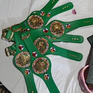 May include: Several green boxing championship belts with gold buckles and circular centerpieces. The centerpieces feature the text "WBC" and images of boxers. The belts are arranged on a white surface.
