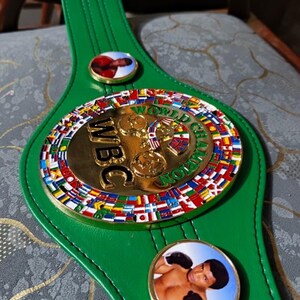 May include: A green leather WBC World Champion boxing belt. The belt features a large gold-colored central medallion with the letters "WBC" and the words "World Champion". The medallion is surrounded by flags of different nations. Two smaller circular medallions with boxer images are on the belt.