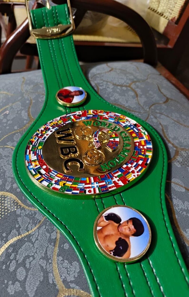 May include: A green leather WBC World Champion boxing belt. The belt features a large gold-colored central medallion with the letters "WBC" and the words "World Champion". The medallion is surrounded by flags of different nations. Two smaller circular medallions with boxer images are on the belt.
