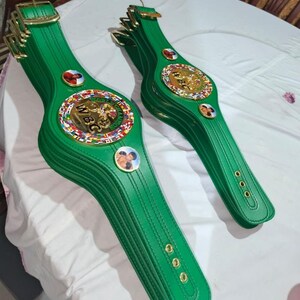 May include: Several green leather championship belts with gold buckles and medallions. The belts feature the WBC logo and images of boxers. The belts are arranged on a white surface.