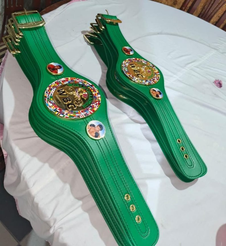 May include: Several green leather championship belts with gold buckles and medallions. The belts feature the WBC logo and images of boxers. The belts are arranged on a white surface.