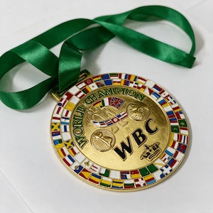 Puede incluir: Una medalla circular dorada de campeón mundial WBC con un diseño central en relieve que incluye el texto WBC, un icono de corona y un gráfico de globo terráqueo. El borde exterior muestra un anillo de banderas internacionales y una cinta verde.