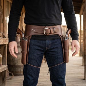 May include: A brown leather gun belt featuring two holsters, each containing a revolver with a wooden grip. The belt includes a metal buckle and leg straps, worn over dark blue denim jeans and a long-sleeved black shirt.