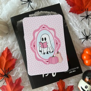 Peut inclure: Une carte rose et blanche sur le thème d'Halloween avec un fantôme de dessin animé lisant un livre, dans un cadre rose. La carte est posée sur un livre noir, entourée de citrouilles, de feuilles et d'araignées décoratives.