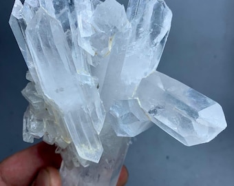 May include: A cluster of clear quartz crystals with multiple points. The crystals are translucent and have a natural, rough texture. The quartz formation is set against a soft, gray background.