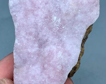 May include: A rough-cut, light pink mineral specimen with a textured surface. The stone has a jagged edge and a brown, earthy base. The overall shape is irregular, with a soft, diffused color.