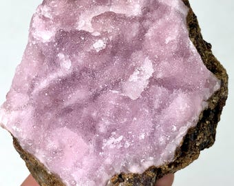 May include: A natural mineral specimen featuring a cluster of light purple crystals. The crystals have a sparkling, granular texture and are set against a dark brown, rough matrix. The overall shape is irregular, showcasing the raw beauty of the stone.