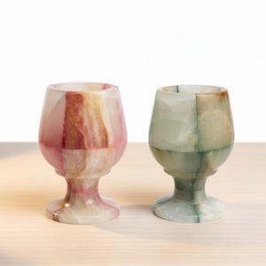 May include: Two decorative onyx goblets. One goblet is pink and white, the other is green and white. The goblets have a rounded bowl shape and sit on a pedestal base. The goblets are on a light-colored wooden surface.