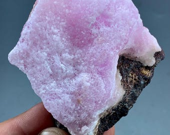 May include: A rough-textured, light purple mineral specimen with a dark brown and white base. The mineral has a crystalline appearance and is held by a hand, showcasing its natural form and color variations.