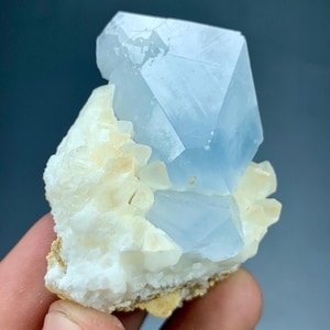 May include: A light blue crystal formation with a large, geometric shape on top of a cluster of smaller, off-white crystals. The crystals are set on a beige base, creating a striking contrast. The image is well-lit, highlighting the crystal's facets.