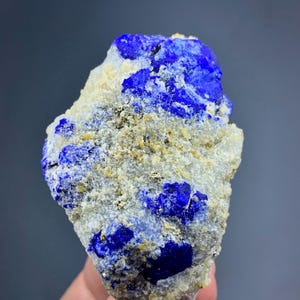 May include: A raw, irregular chunk of lapis lazuli mineral featuring deep blue crystalline sections embedded within a rough, light gray and beige matrix. Small, metallic gold-colored pyrite flecks are scattered across the textured surface.