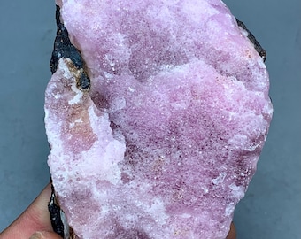 May include: A rough-hewn, natural mineral specimen. The rock features a large, irregular shape with a prominent, textured surface. The color is a soft, dusty rose, with areas of darker, almost black, mineral deposits along the edges.