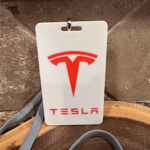 May include: White rectangular Tesla ID badge with a red logo and the word "TESLA" in red. The badge is attached to a gray lanyard. The background is a textured brown surface.