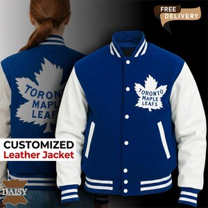 Op de afbeelding: Een blauw-witte varsity-jas met het logo van de Toronto Maple Leafs op de borst en rug. De jas heeft een sluiting met drukknopen, witte mouwen en gestreepte geribbelde boorden bij de mouwen en de kraag met de tekst Toronto Maple Leafs.