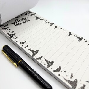 May include: A white notepad with the words "Witchy Notes" in black, decorated with witch hats, bats, moons, and stars. A black pen with a gold clip rests on the notepad. The notepad has lined pages.