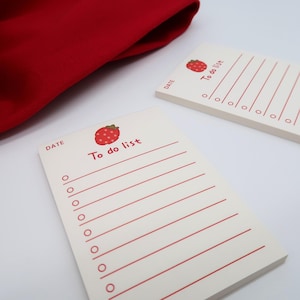 Puede incluir: Dos blocs de notas crema con listas de tareas, líneas rojas y una ilustración de fresa. El texto "To do list" está escrito en rojo. Un tejido rojo está en el fondo.