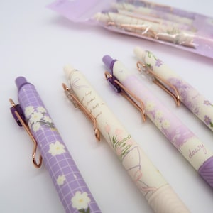 Puede incluir: Cuatro bolígrafos morados y blancos con diseños florales y detalles dorados. Los bolígrafos tienen un clip y un cuerpo transparente. El texto en los bolígrafos dice "your leaf, sweet text in summer, daisy".