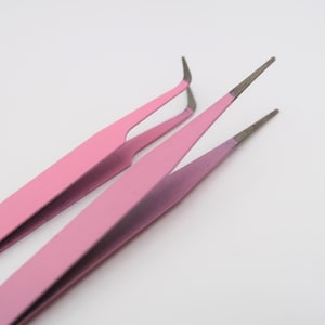 May include: Two pink tweezers with slanted tips. The tweezers are made of metal and have a matte finish.