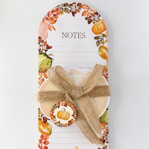 Può includere: Un blocco note decorativo a tema autunnale. Il blocco note ha uno sfondo bianco con linee per scrivere e la parola "NOTES" in alto. È adornato con un disegno ad acquerello di zucche e fogliame autunnale. È attaccato un tag rotondo con l'immagine di una zucca.