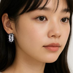 Puede incluir: Primer plano de una persona que lleva un pendiente en blanco y negro. El pendiente tiene forma cuadrada con un borde dentado. El pendiente tiene texto japonés en blanco.